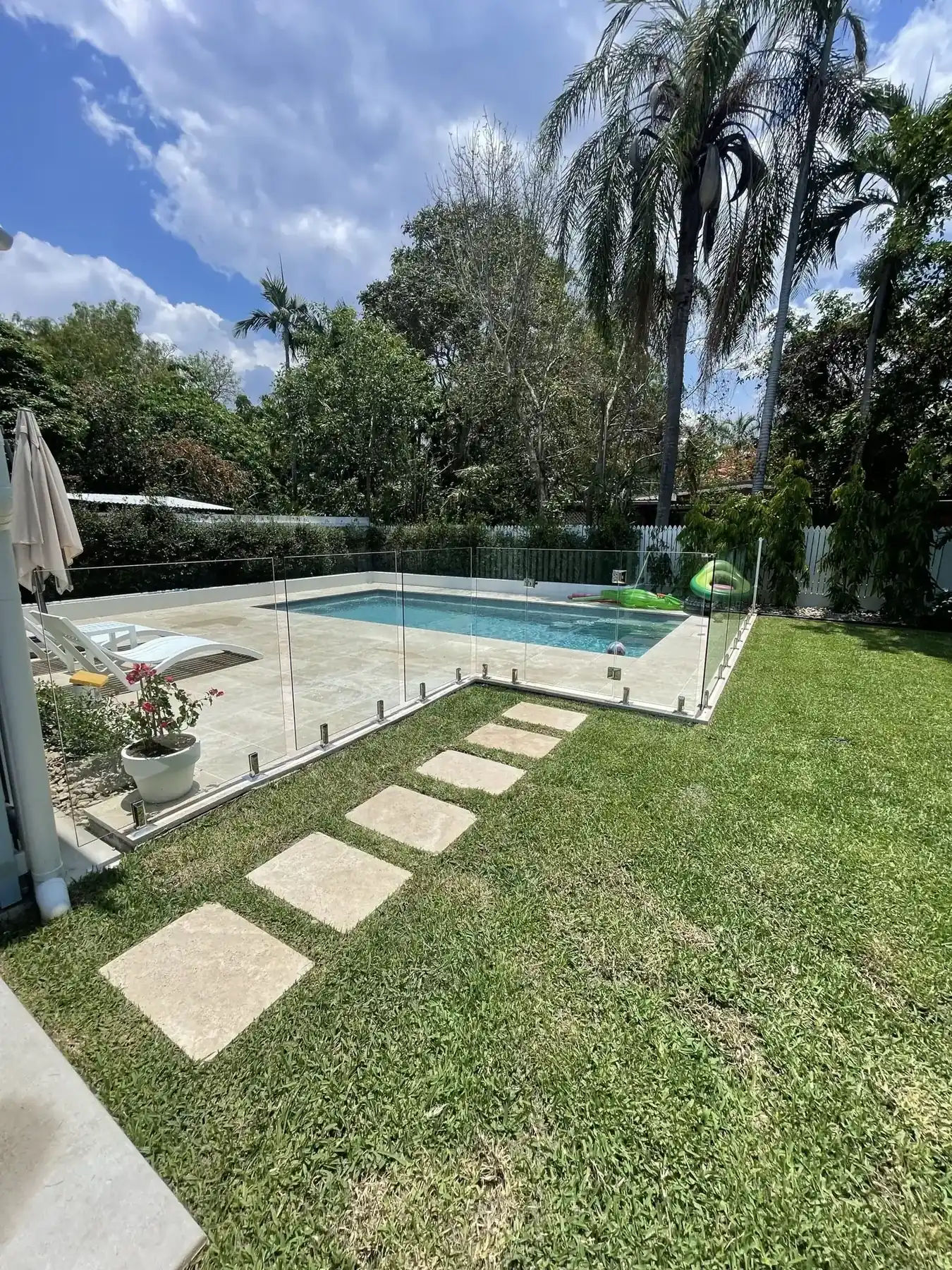 Glass pool fence and stepping-stone path beside a home pool with palm trees